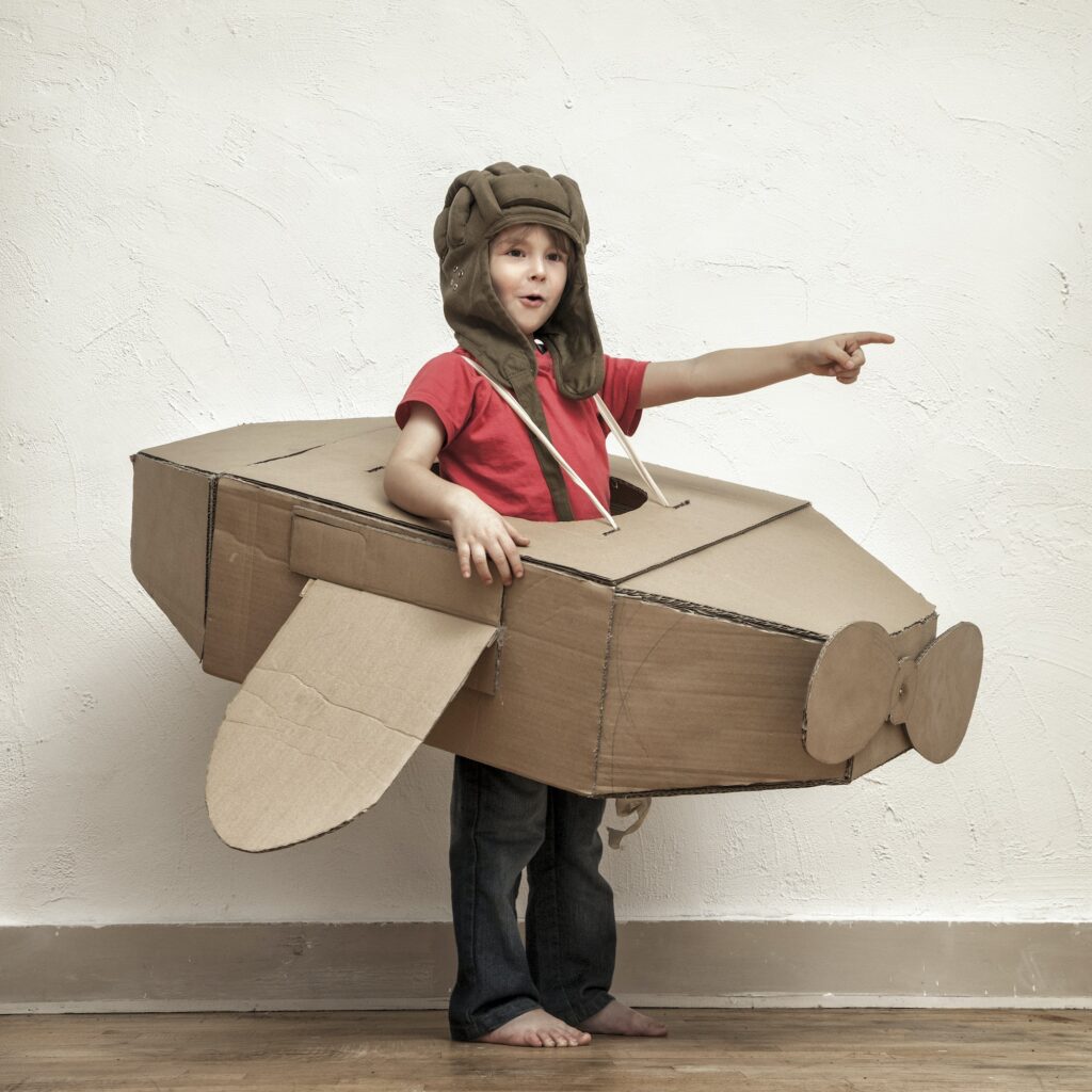 Little boy with pilot hat and cardboard box aeroplane showing at something